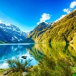 Tranquil alpine lake in snowy valley with lush greenery, reflecting clear blue skies.