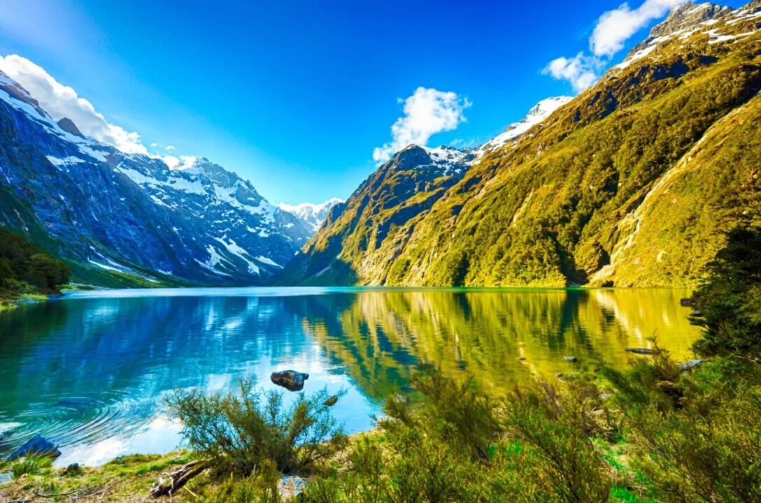 Tranquil alpine lake in snowy valley with lush greenery, reflecting clear blue skies.