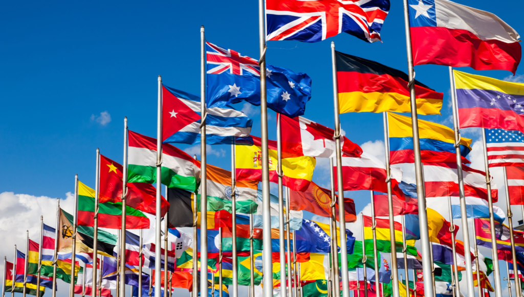 A vibrant display of various national flags fluttering on poles against a blue sky with fluffy clouds.