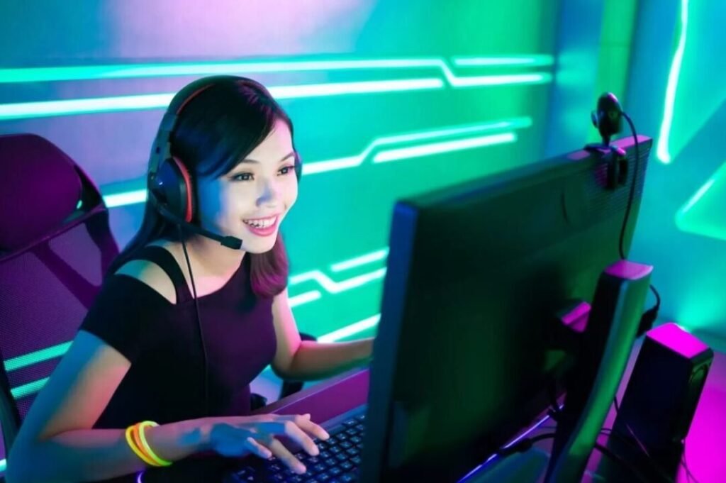 Young Asian woman smiling and using a computer with headphones in a neon-lit gaming setup.