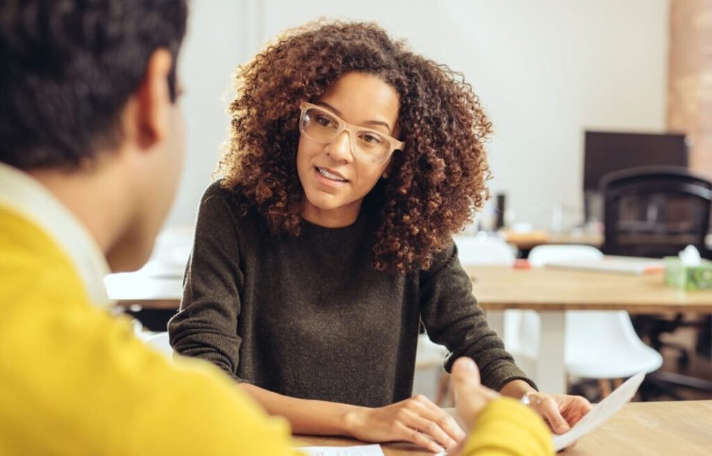 Woman with curly hair and glasses in a discussion with a person across the table in an office environment.