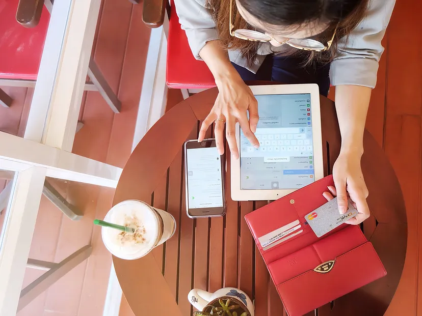 Overhead view of a person at a wooden table with a tablet, smartphone, red wallet, and a cold beverage with a green straw. The person is interacting with both the tablet and the smartphone.