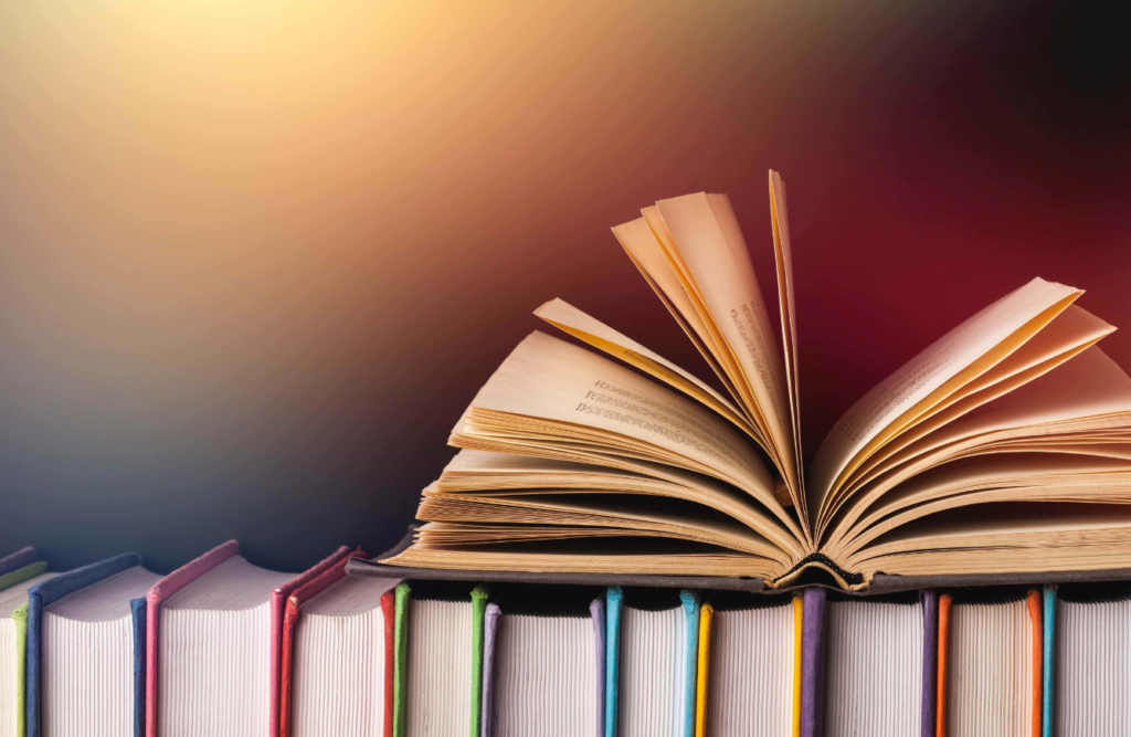 An open book with pages fanned out on top of a row of colorful, upright books against a dark background with a warm glow.