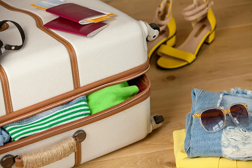 A partially packed suitcase with clothing items and travel documents, a pair of high heel sandals, and folded jeans with sunglasses on top, suggesting travel preparations.