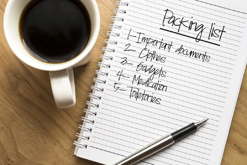 A notepad with "Packing list" written at the top and a list below that includes "1- Important documents, 2- Clothes, 3- Gadgets, 4- Medication, 5- Toiletries", next to a cup of black coffee and a pen on a wooden surface.