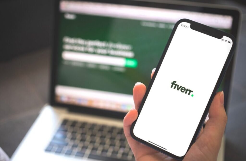 A person holding a smartphone with the Fiverr logo on the screen, with a blurred background showing a laptop with text and a green banner.