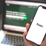 Freelancer using Fiverr on smartphone in modern workspace.