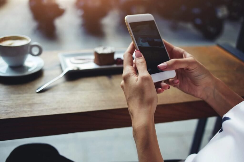 Person using a smartphone at a cafe table with a coffee cup and dessert in the background.