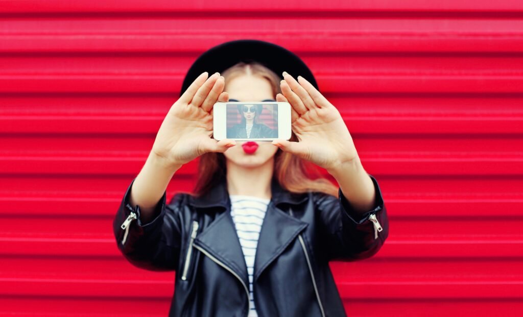 A woman holding a smartphone in front of her face displaying a selfie, stylishly posing against a red corrugated background.