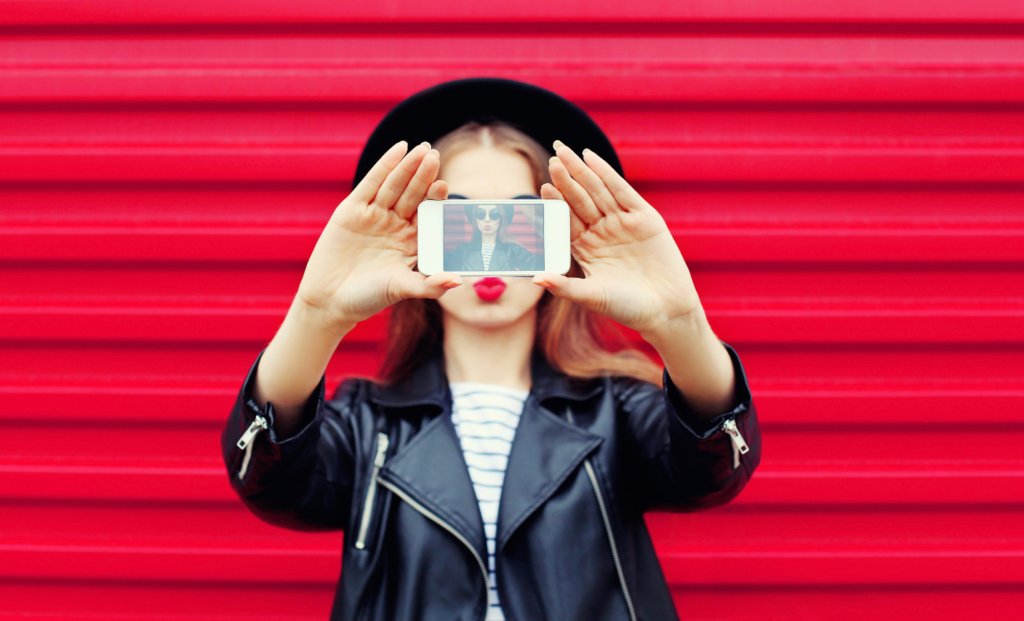 A woman holding a smartphone in front of her face displaying a selfie, stylishly posing against a red corrugated background.