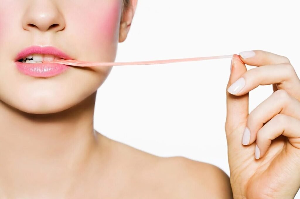 A close-up photo of a woman pulling a pink chewing gum with her teeth and fingers, showing her pink lips and rosy cheeks against a white background.