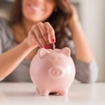 Woman saving money by depositing coin into pink piggy bank on table.