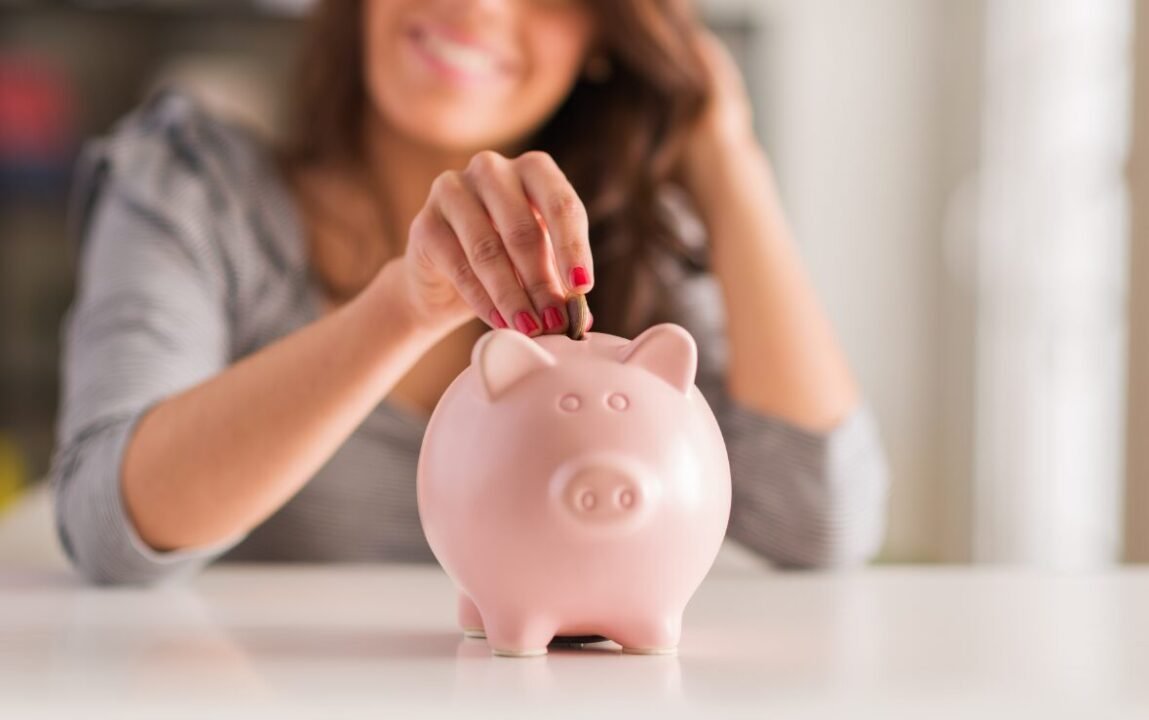 Woman saving money by depositing coin into pink piggy bank on table.