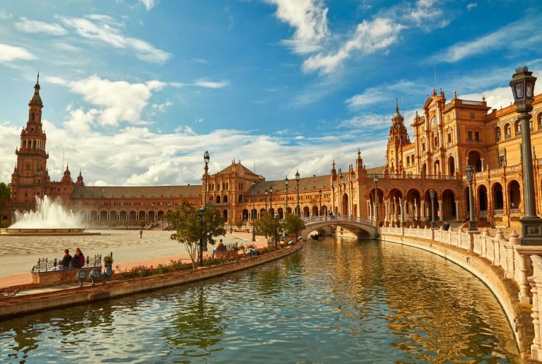 Plaza de España in Seville with a prominent building featuring Renaissance/Neo-Moorish architecture, a canal with a bridge, and a fountain under a cloud-streaked sky.