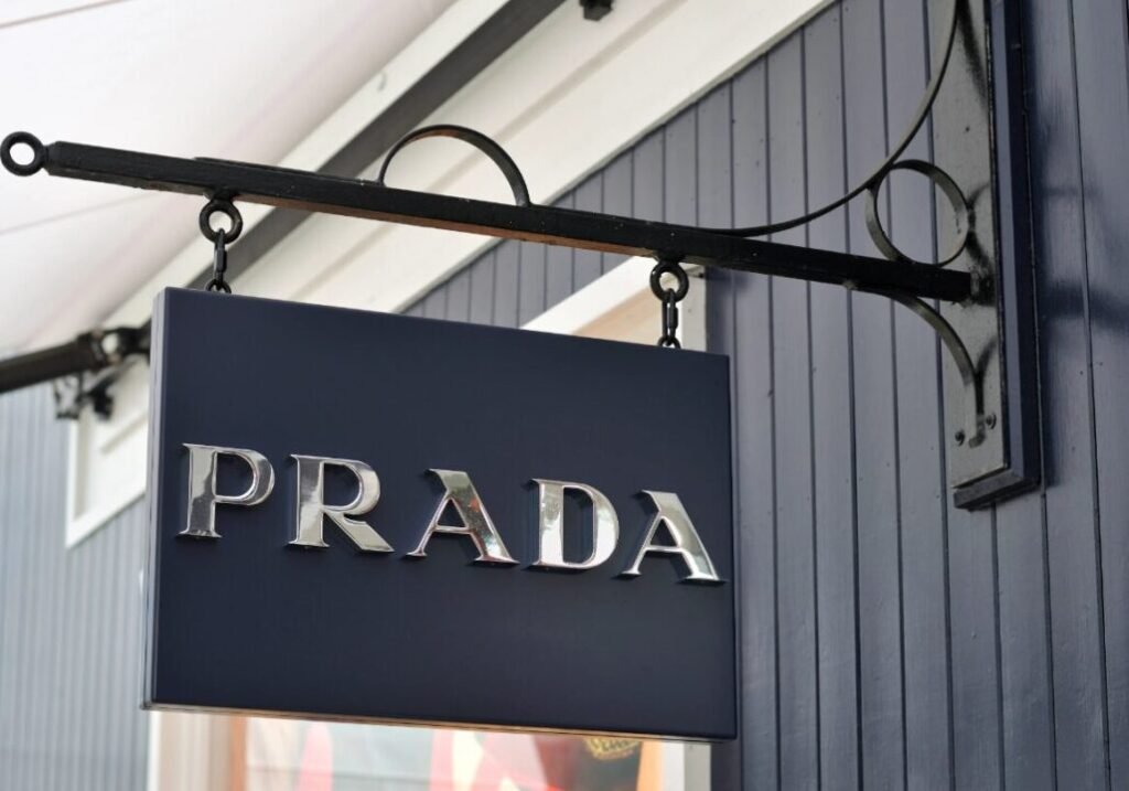 Prada brand sign hanging on black metal bracket outside a building with gray wooden siding.