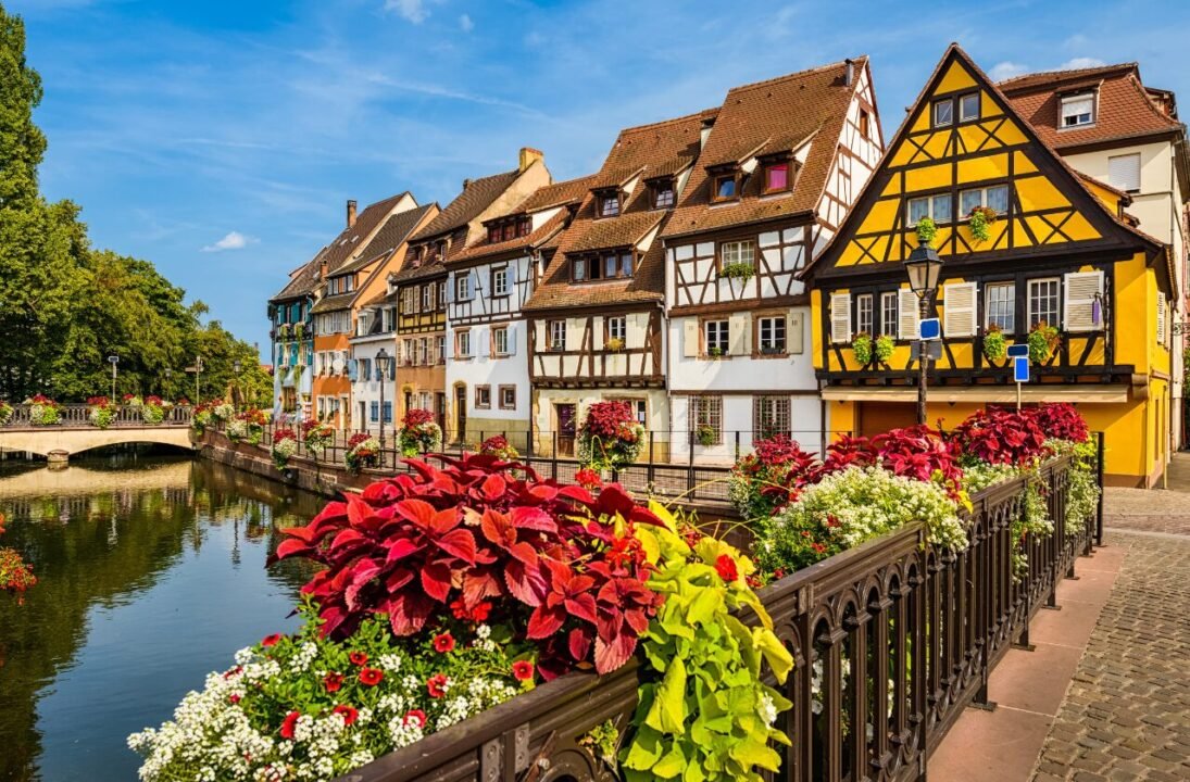 Quaint half-timbered houses along a canal, adorned with colorful flowers in full bloom, under a clear blue sky.