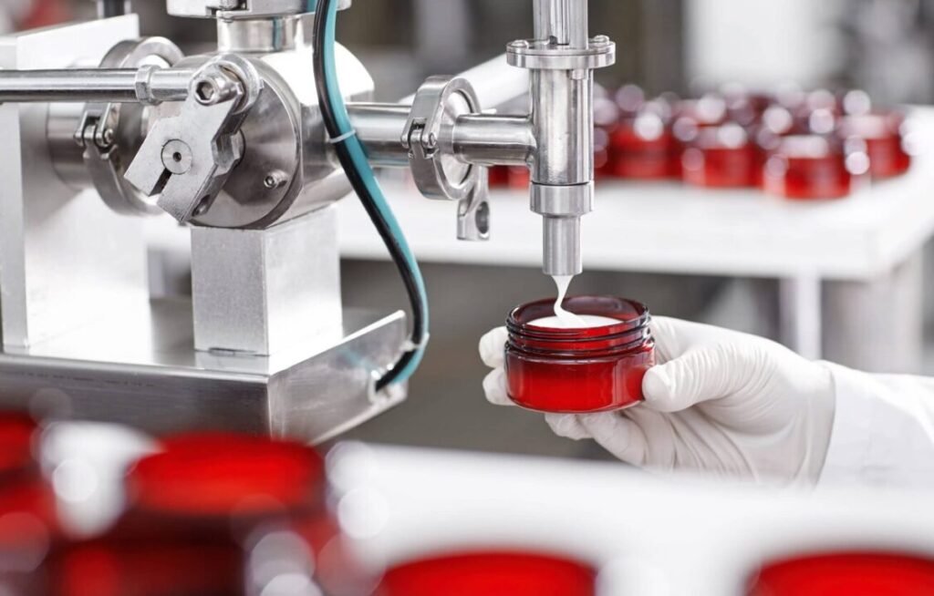 A person in lab gloves filling red cosmetic jars with a white cream from an industrial dispensing machine in a manufacturing facility.