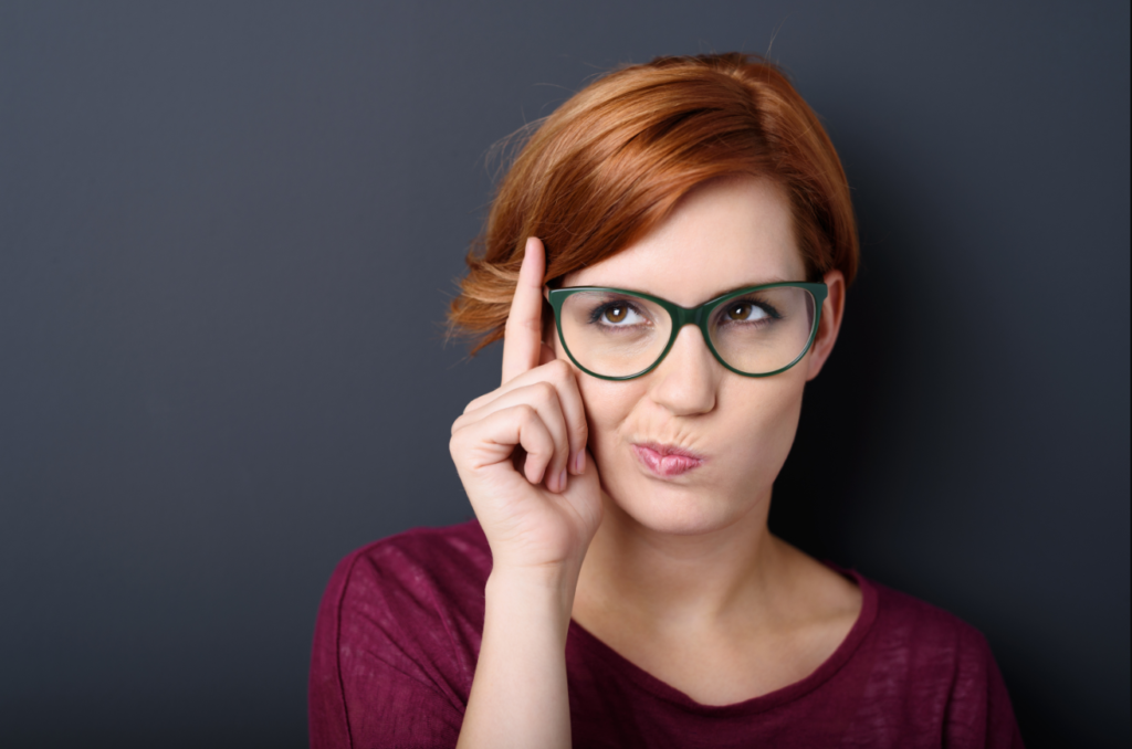 A woman with red hair and green glasses raising her index finger to her temple, making a pensive face against a dark gray background.