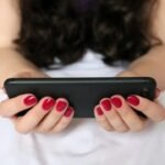 Person holding smartphone with red nail polish on bed, engaging with modern technology.
