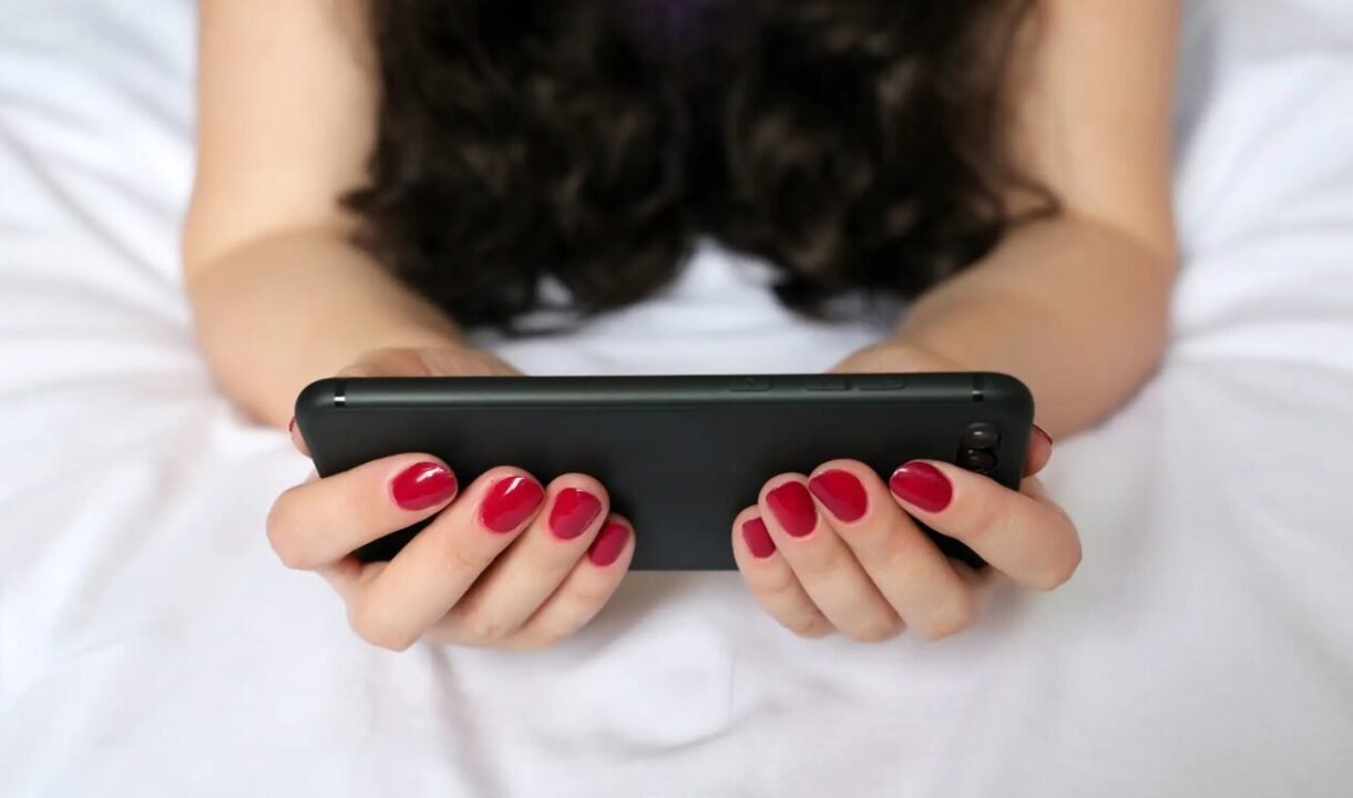 Person holding smartphone with red nail polish on bed, engaging with modern technology.