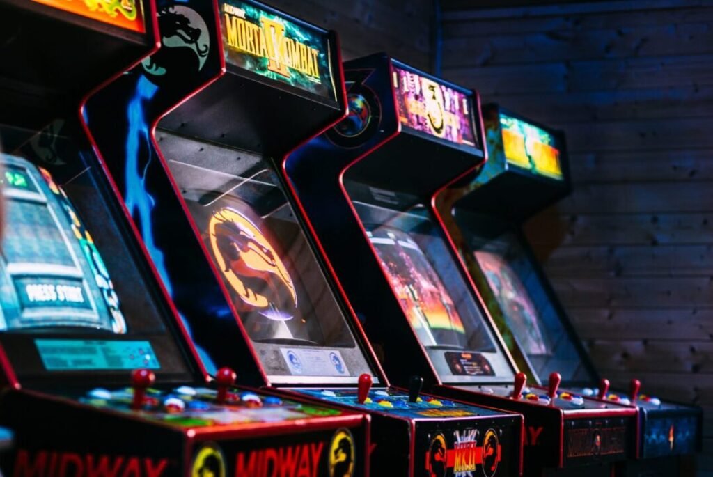 A row of retro arcade machines, including Mortal Kombat, brightly lit in a dim room with wooden walls.