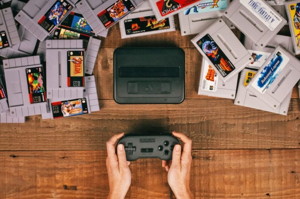 A person holding a classic video game controller with numerous retro game cartridges and a gaming console scattered on a wooden surface.