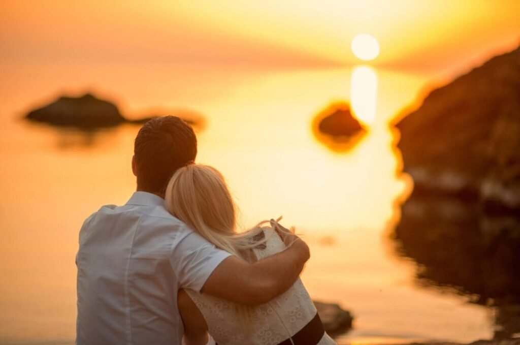 Loving couple watching sunset together by tranquil sea, embracing in warm, romantic moment.