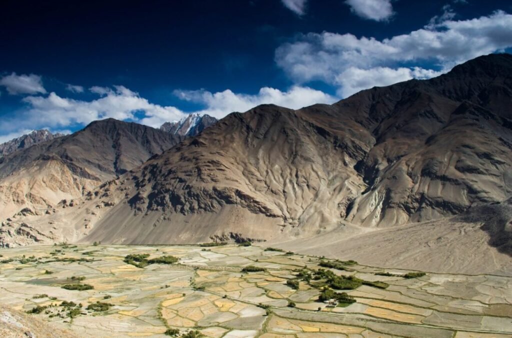 Alt text: A rugged landscape featuring a valley with patchwork fields bordered by desolate towering mountains under a blue sky with scattered clouds.