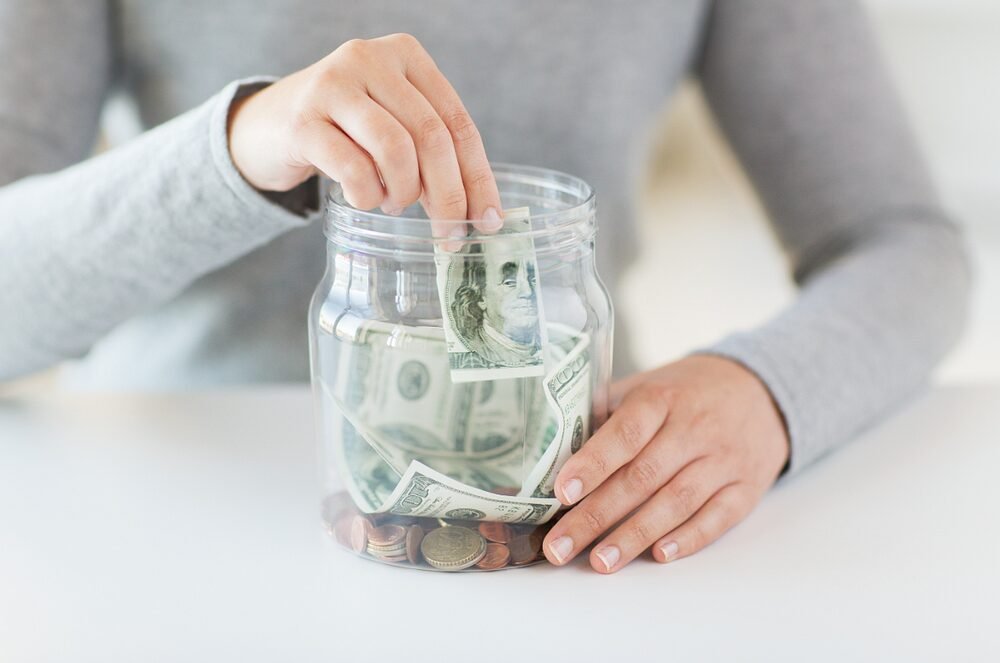 Person saving money by adding a dollar bill to a jar of mixed U.S. currency.