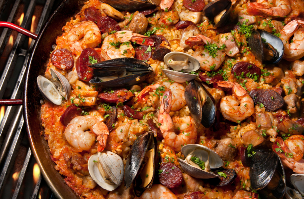 A close-up image of a seafood paella with shrimp, mussels, clams, and chorizo, garnished with parsley, cooked in a large pan on a grill.