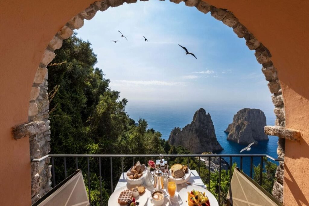 A breakfast table set with food on a balcony with a seaside view framed by an arched stone entryway, with nearby seagulls flying and rock formations projecting from the sea.