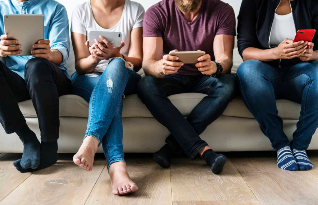 Four people sitting side by side on a couch, each engrossed in their own tablets and smartphones, showcasing a mix of casual footwear and bare feet.