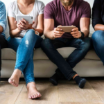 Modern social scene: People on couch immersed in devices, casual attire, varied footwear.