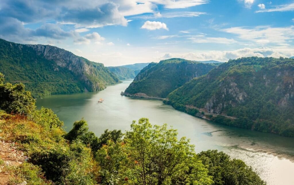 Scenic view of a winding river flowing between tall forested cliffs under a partly cloudy sky, with a barge visible on the water.