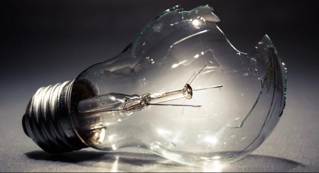 A broken clear glass light bulb with visible filament and base, set against a grey background with light reflections on the shattered pieces.