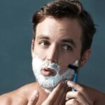 Man shaving with razor, focused expression, white foam on face.