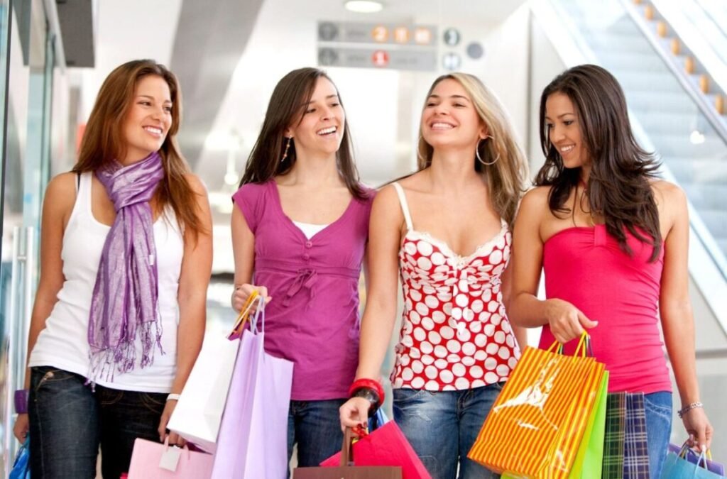 Four women walking and laughing in a shopping mall, each carrying colorful shopping bags.