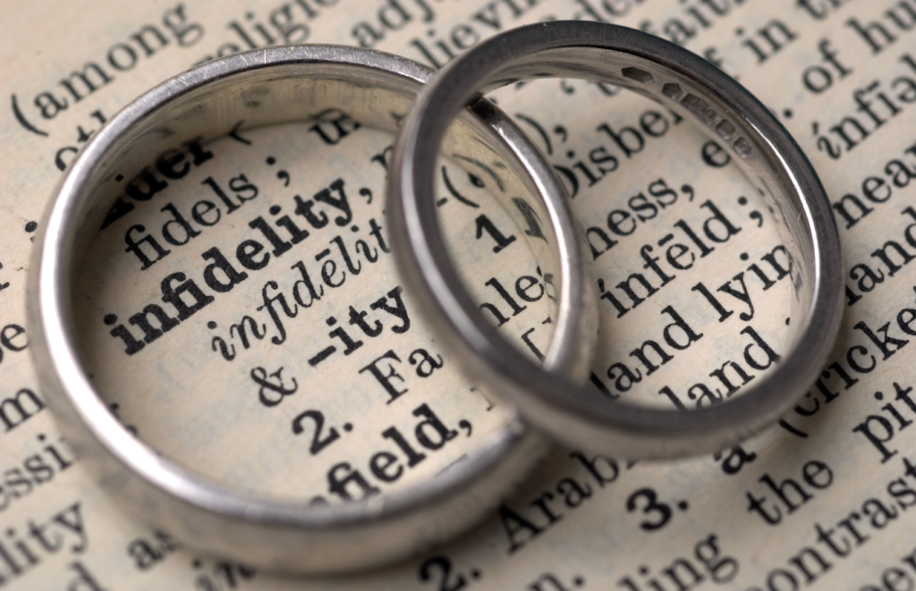 Two silver wedding rings overlapping on top of a dictionary page with the word 'infidelity' visible through the rings.