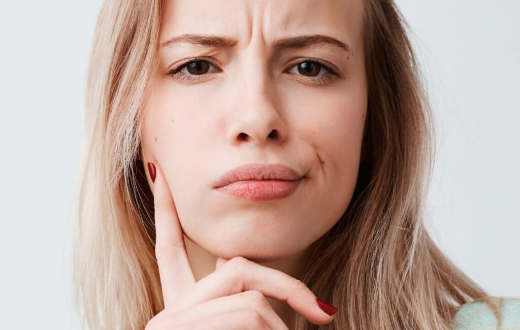 Close-up of a skeptical young woman with blonde hair, touching her chin and frowning, isolated on a light background.