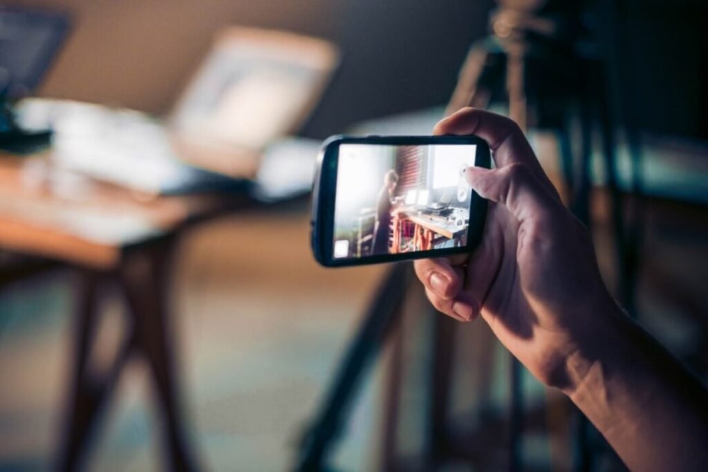 Person holding a smartphone taking a photo of another person working in an office, visible through the phone's screen.