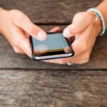 Persons hands using smartphone on wooden table, engaging with colorful screen content.