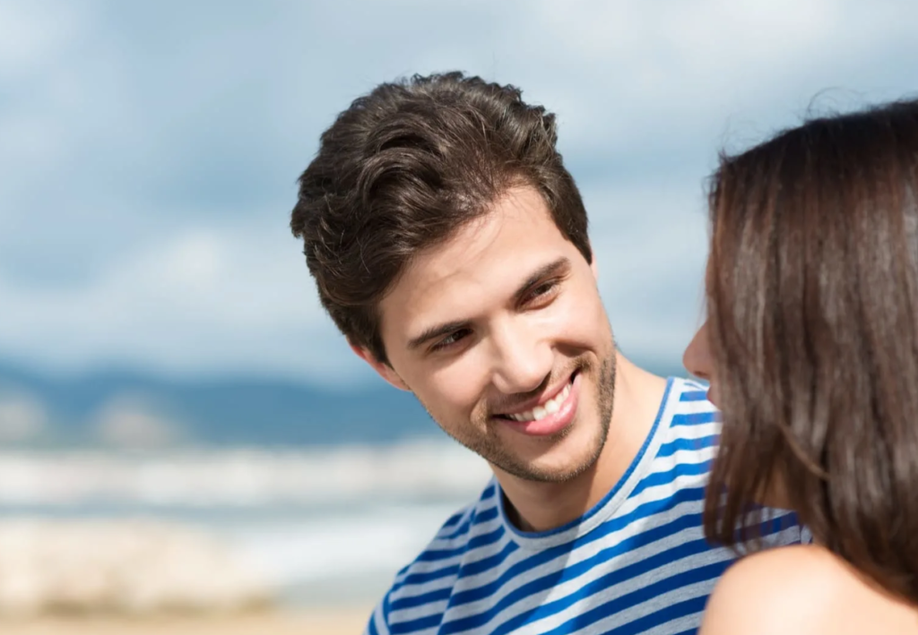 A smiling man in a blue and white striped shirt looking at a person with dark hair, with a blurred beach and sky in the background.