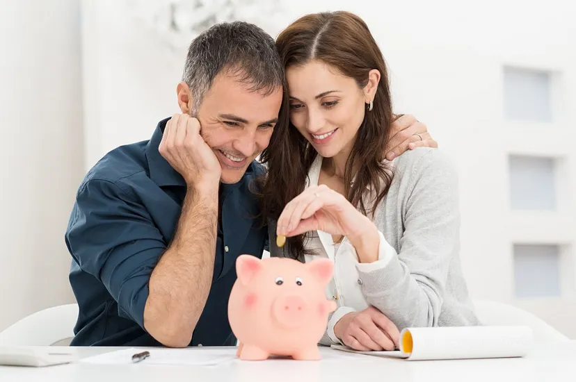 A smiling man and woman inserting a coin into a piggy bank on a table with papers rolled up next to them.