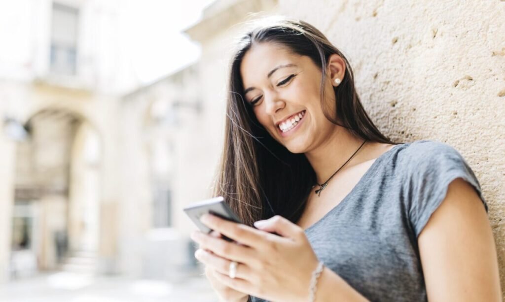 Young woman smiling and using smartphone against a textured stone wall in a sunny outdoor setting.