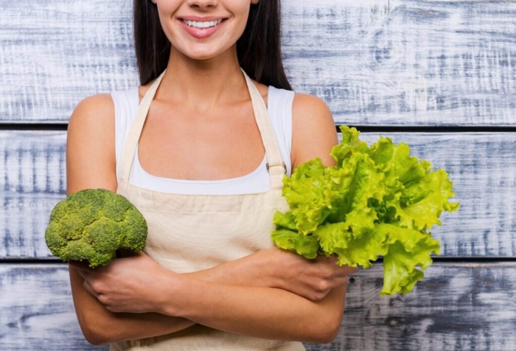 A smiling woman wearing an apron holds a broccoli in one hand and a bunch of lettuce in the other, standing before a blue wooden background.