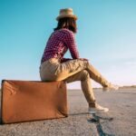 Contemplative traveler on vintage suitcase gazes into open road under clear blue sky.