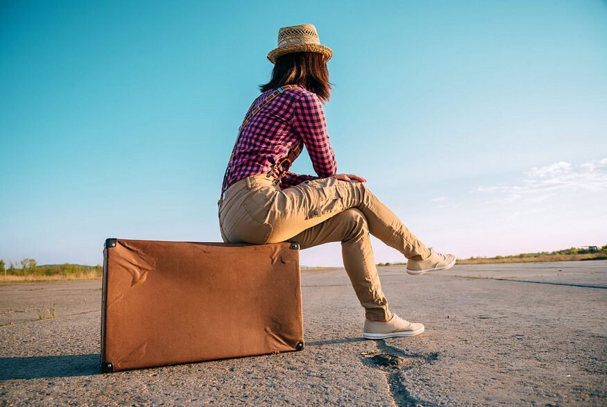 Contemplative traveler on vintage suitcase gazes into open road under clear blue sky.