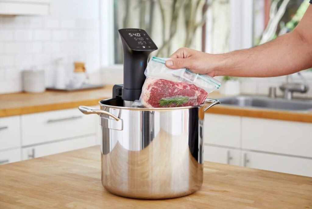 Person placing a vacuum-sealed bag with steak and herbs into a sous vide cooker in a kitchen.