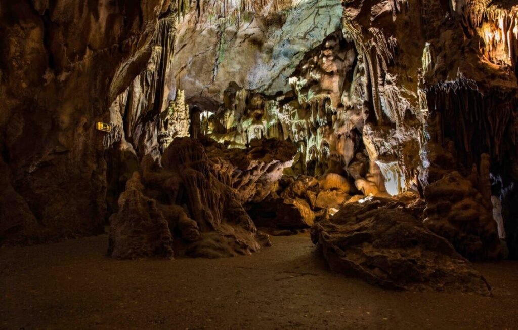 A dimly lit cavern with various stalactites and stalagmites, and a dirt floor scattered with rocks.