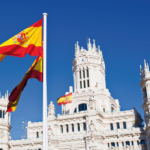Spanish flag waving in front of majestic Neo-Classical palace under clear blue sky.
